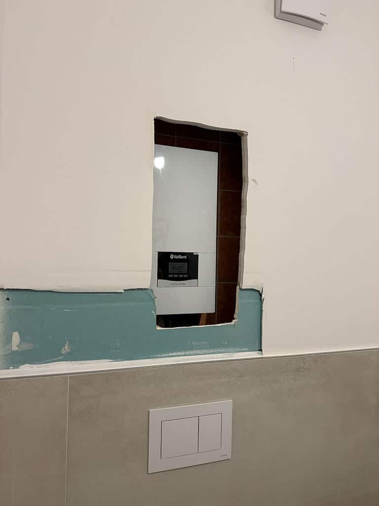 Badsanierung mit Revisionsöffnung hinter WC-Vorwandinstallation Revisionsöffnung in einer WC-Vorwandinstallation während einer Badsanierung mit sichtbarer Vaillant-Heizungssteuerung im Hintergrund.